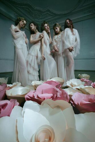 Des mannequins portent des robes longues en tulle blanc, entourées de grandes fleurs roses et blanches, dans un environnement minimaliste évoquant une ambiance romantique et vaporeuse.