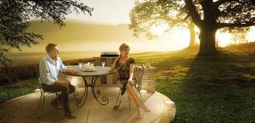 Deux mannequins élégamment habillés sont assis à une table extérieure au lever du soleil, créant une ambiance chic et sophistiquée dans un décor naturel.