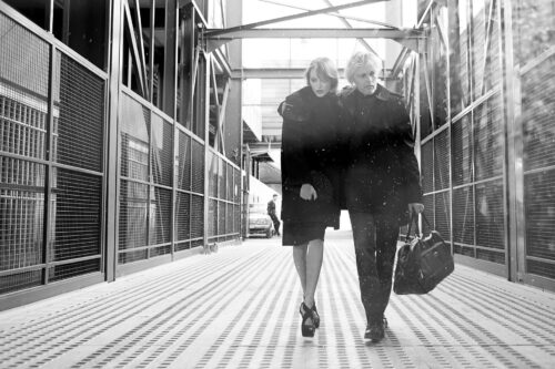 Deux mannequins marchent sur une passerelle industrielle, habillés élégamment en noir chic, évoquant un style contemporain sophistiqué et citadin.