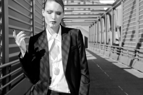 Mannequin en costume noir élégant, chemise blanche, cravate fine, maquillage marqué, pose sophistiquée sur un pont métallique, style androgynique et moderne, ambiance urbaine.