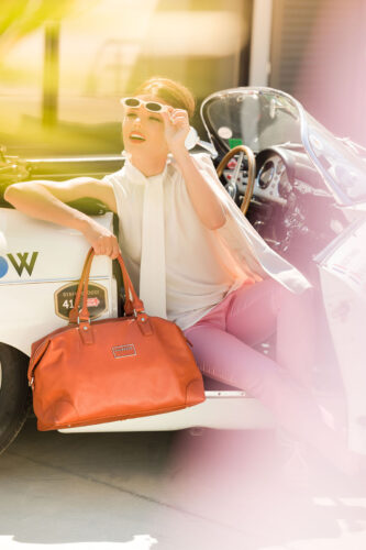 Mannequin élégant assis dans une voiture vintage, portant un pantalon rose, un haut blanc et des lunettes de soleil, avec un grand sac à main orange sophistiqué.