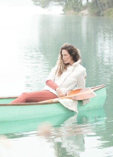 mannequin porte une doudoune blanche élégante et un pantalon rouge, assise dans une barque verte sur un lac paisible, évoquant une mode hivernale décontractée.