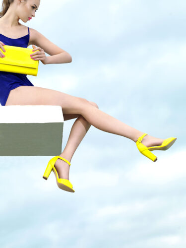 Mannequin en robe bleue, accessoirisée de chaussures à talons et pochette jaunes, pose élégante, style contemporain sur fond de ciel bleu.