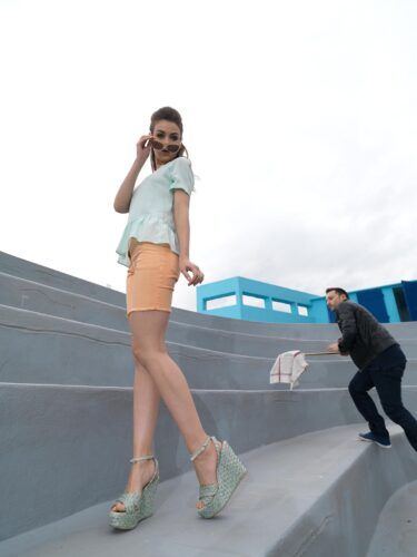 Mannequin portant un short pêche, chemisier bleu clair et sandales compensées imprimées, posant sur des marches grises avec élégance; style urbain et contemporain.