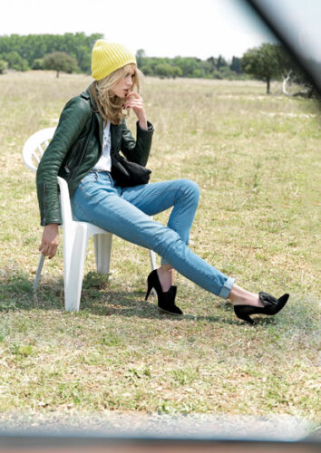 Mannequin assis, portant un jean, blouson en cuir vert, t-shirt blanc, talons noirs et bonnet jaune, dans un style décontracté-chic extérieur.