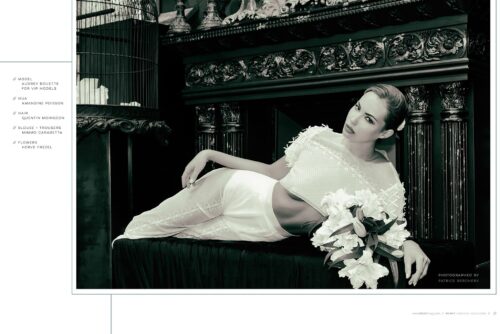 Mannequin posant élégamment dans une tenue blanche sophistiquée, mise en scène sur un canapé sombre, entourée de fleurs et d’éléments décoratifs classiques.