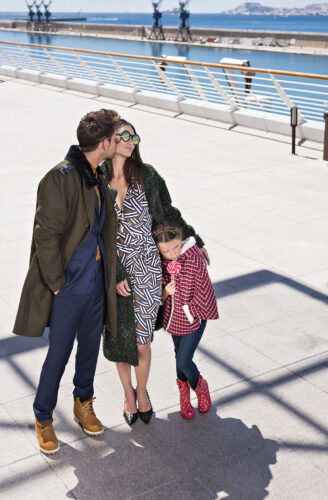 Des mannequins élégamment habillés posent en extérieur ; l'homme porte un manteau long, la femme une robe imprimée, l'enfant un manteau rayé et des bottes rouges.