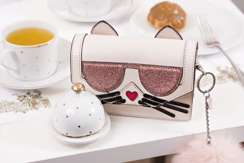 Sac à main fantaisie en forme de chat, détails pailletés et graphiques, accessoire mode ludique idéal pour un look original et tendance, posé sur une table avec tasse de thé.