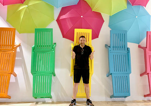 Installation scénographique colorée avec chaises longues et grands parapluies accrochés au mur, évoquant une animation estivale ou une décoration de vitrine dans un espace public.