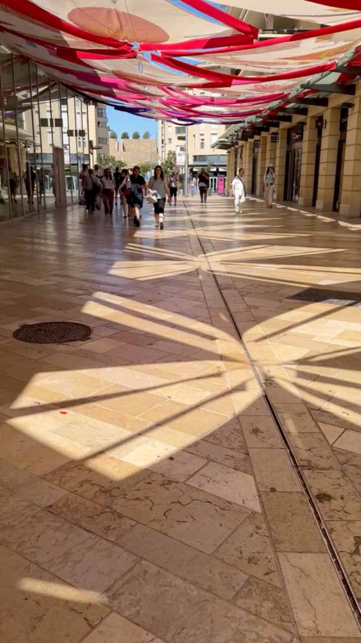 Des motifs géométriques lumineux sont projetés au sol, résultant d’installations artistiques en hauteur, créant une scénographie dynamique dans une galerie commerçante piétonne.