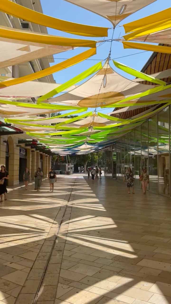 Des installations scénographiques de voiles colorées flottent au-dessus d’une rue piétonne, projetant des ombres dynamiques sur le sol, créant une ambiance urbaine estivale et artistique.