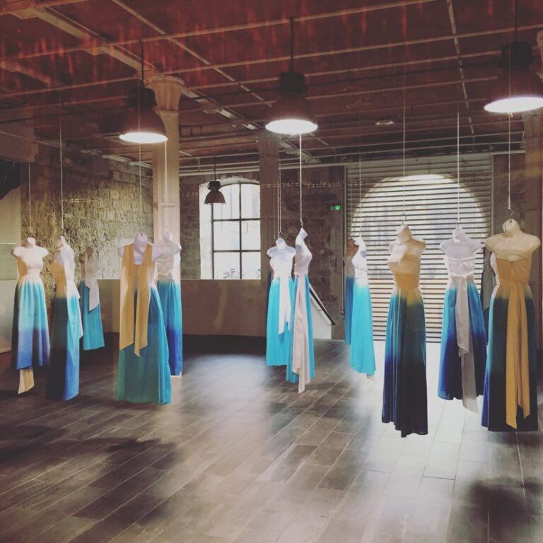Installation scénographique de robes suspendues sur des mannequins, éclairée par des spots dans un espace industriel, évoquant une vitrine artistique ou une animation de centre commercial.