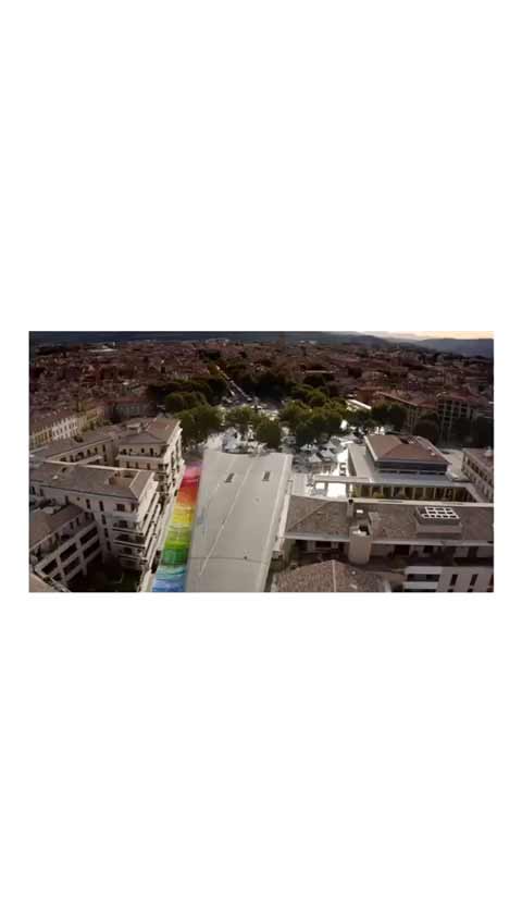 Vue aérienne d'une installation scénographique urbaine avec une large bande colorée arc-en-ciel traversant une place, intégrée à l’environnement architectural contemporain.