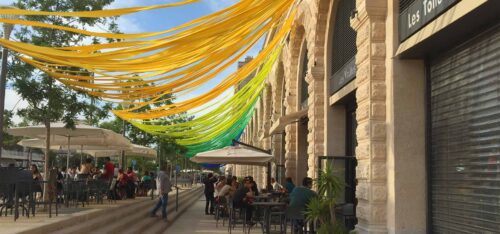 Installation scénographique de rubans colorés flottant au-dessus d'une terrasse de café, devant une façade en pierre, créant une ambiance festive dans l’espace urbain.