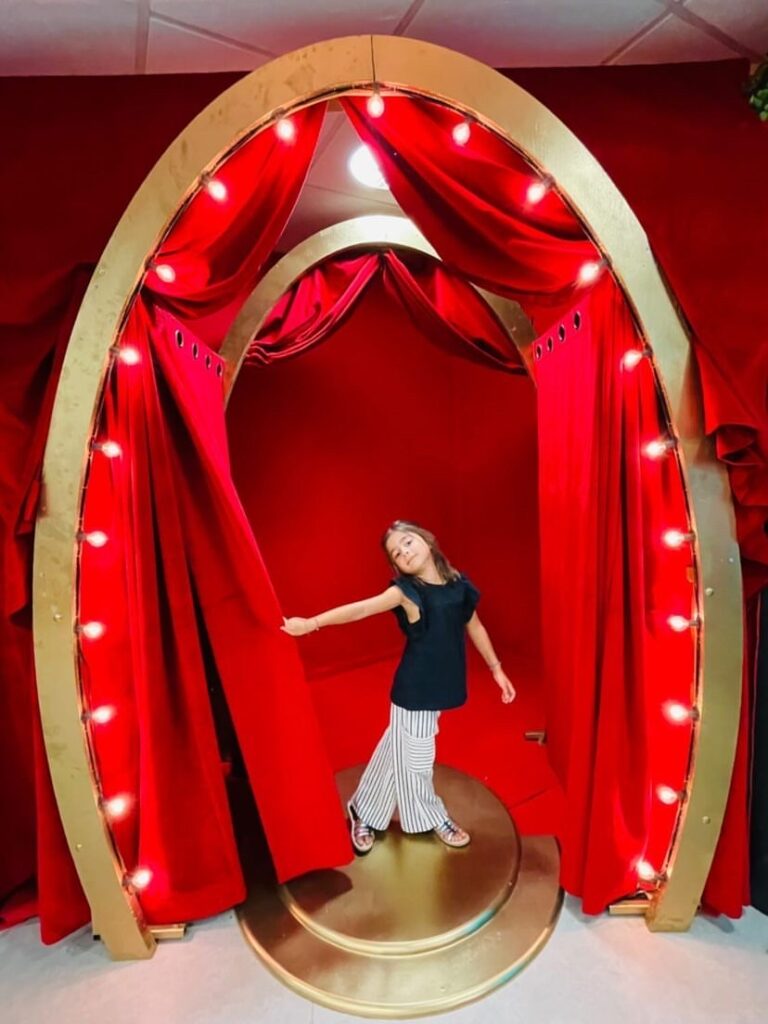 Installation scénique composée d'une arche ovale dorée, entourée de rideaux rouges lumineux, créant l'illusion d'une entrée théâtrale, avec une personne posant de façon élégante.