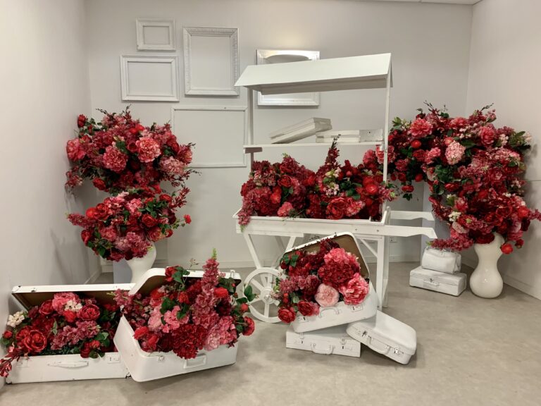 Installation scénographique de vitrines composée d’un chariot, de caisses et de cadres blancs, ornés de grands bouquets de fleurs rouges et roses, créant une ambiance élégante et florale.