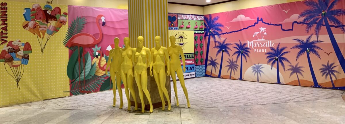 Groupe de mannequins jaunes installés autour d’une colonne, devant des décors colorés à motifs tropicaux et graphiques évoquant une ambiance estivale en vitrine.
