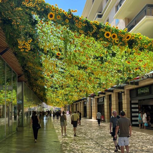 Installation scénographique florale suspendue recouvrant la rue, composée de tournesols et feuillages, créant une ambiance immersive et ombragée pour les passants et les vitrines commerçantes.
