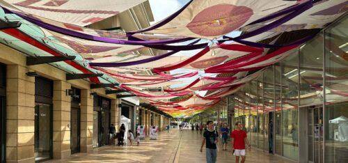 Installation scénographique de tissus colorés suspendus créant une couverture décorative au-dessus d’une rue commerçante piétonne, apportant ombre et dynamisme à l’espace public.
