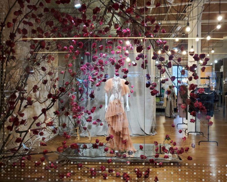 Installation scénographique de vitrine : mannequin vêtu d’une robe rose pâle, entouré de branches décoratives fleuries et de guirlandes lumineuses, créant une ambiance poétique et immersive.