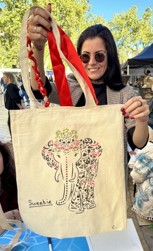 Présentation scénique d’un sac illustré, motifs d’éléphant artistiques en vitrine, décoré de rubans rouges, ambiance marché en arrière-plan.