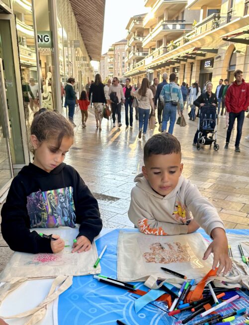 Deux enfants participent à une installation artistique sur un trottoir animé, dessinant avec des fournitures créatives, encadrés par la scénographie vivante d’une rue commerçante.