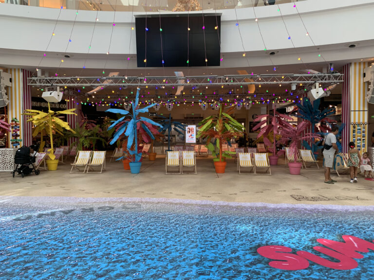 Installations scénographiques colorées en forme de palmiers, disposées devant une vitrine décorée de guirlandes lumineuses, avec des chaises longues et une piscine au premier plan.