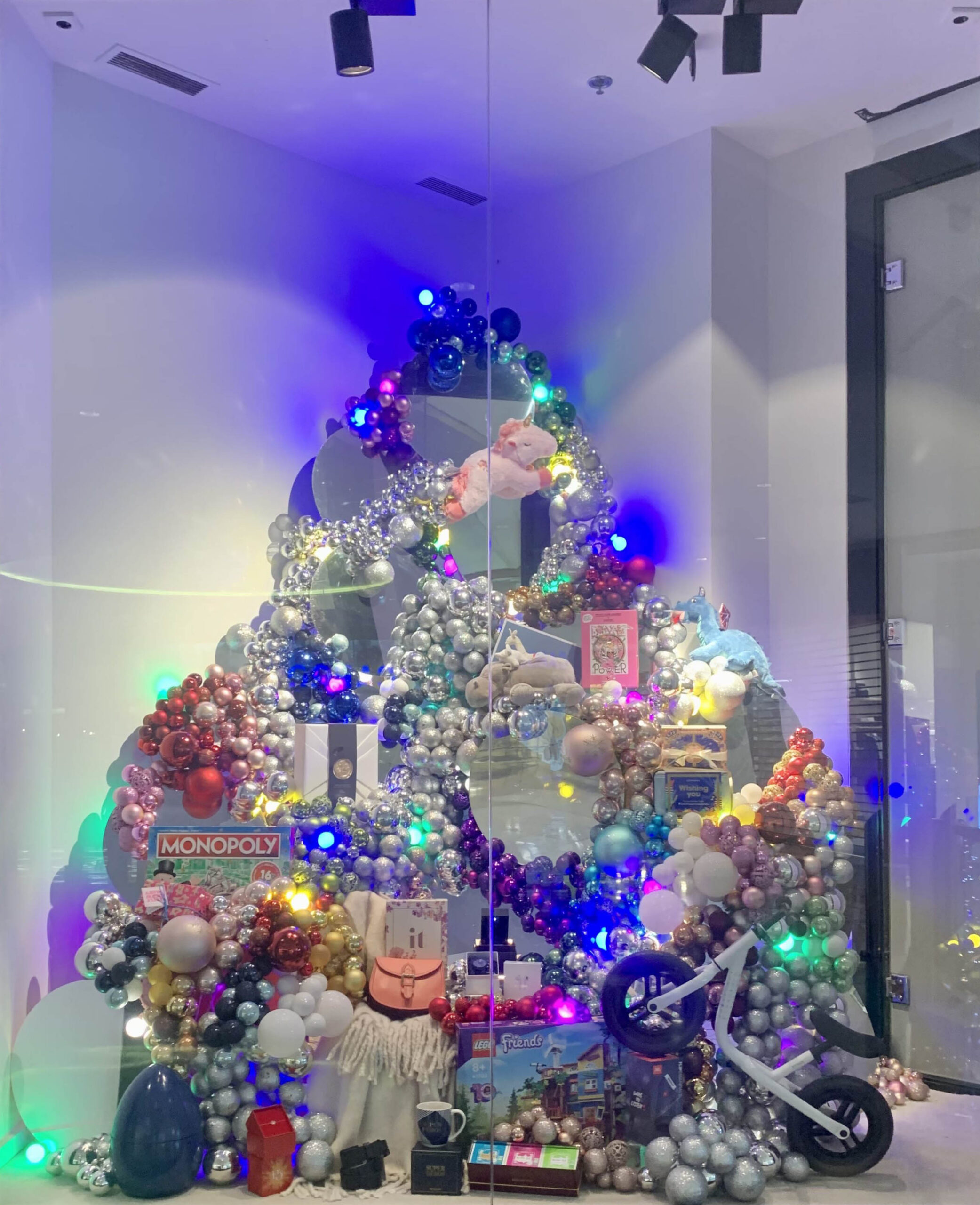 Installation scénographique représentant un sapin stylisé, composé de nombreux objets, cadeaux, guirlandes lumineuses et décorations colorées, exposée derrière une vitrine dans un espace contemporain.