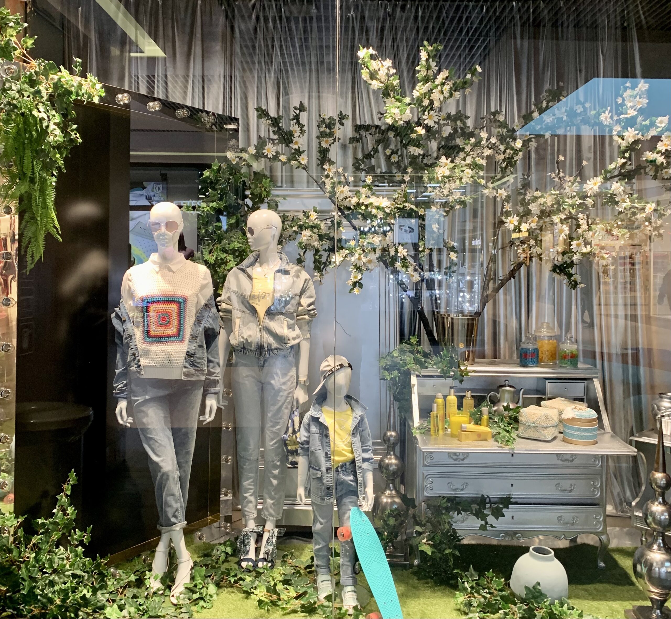 Installation scénographique avec mannequins vêtus, décor végétal, arbres artificiels, éléments de jardin et présentoir de fruits, simulant une scène extérieure en vitrine de magasin.