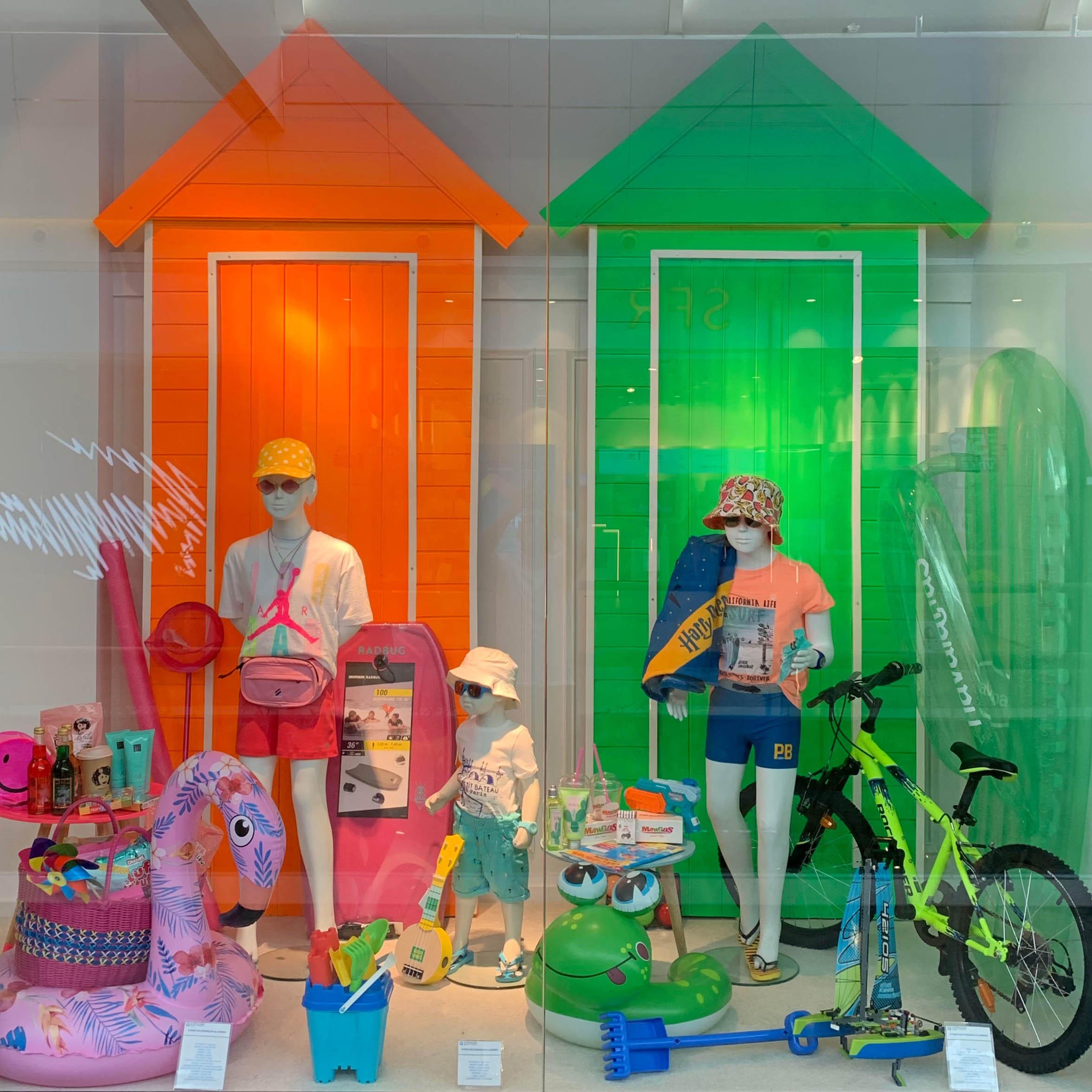 Installation scénographique de vitrine, mannequins enfants vêtus d’accessoires estivaux devant deux cabines colorées, décor complété de jouets, vélo et objets de plage clairs et colorés.