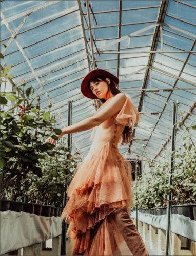 Mannequin portant une robe en tulle rose délicate et un chapeau bordeaux, posant dans une serre lumineuse au style bohème et élégant.