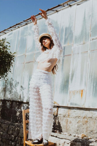 Mannequin portant un ensemble blanc transparent à volants, pose élégante dehors, accessoirisé d’une casquette beige, style contemporain avec influence bohème et accents texturés.