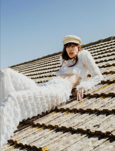 Mannequin posant sur un toit en tenue blanche texturée et élégante, assortie d’une casquette crème, dans une ambiance moderne et décontractée.