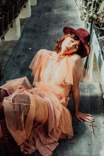 Mannequin posant en robe en tulle rose pâle, chapeau bordeaux élégant, assise de façon décontractée, ambiance lumineuse, accent mis sur la fluidité des matières et le style bohème chic.