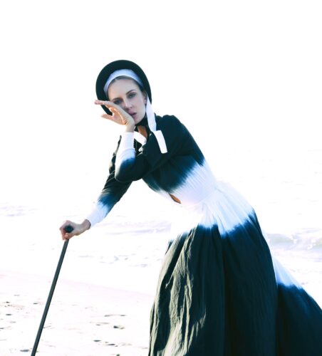 Mannequin en robe longue noire et blanche, style victorien, coiffé d’un bonnet noir, posant avec élégance sur une plage, accessoirisé d’une canne sophistiquée.