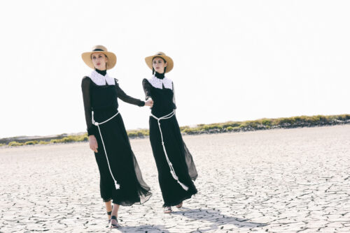 Deux mannequins portent des robes noires longues avec col blanc, ceintures cordon, chapeaux de paille ; look épuré et contrasté, style inspiré du minimalisme vintage.