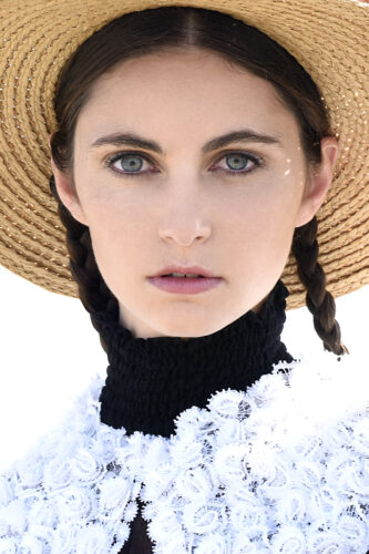 Mannequin portant un chapeau de paille à larges bords, deux tresses, col haut noir, vêtue d'une blouse blanche en dentelle, style champêtre sophistiqué.