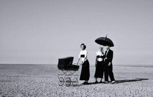 Des mannequins posent en tenues élégantes noir et blanc sur une plage déserte, l’un tenant un parapluie, l’autre poussant une poussette rétro, ambiance mode sophistiquée.