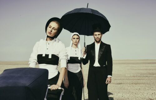 Trois mannequins en tenues inspirées du style amish, noir et blanc, posent dans un paysage aride, l’un tenant un parapluie bleu foncé.
