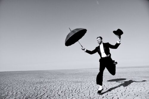 mannequin masculin en costume élégant, sautant sur un sol aride, tient un parapluie et un chapeau, style classique et sophistiqué, ambiance noir et blanc.