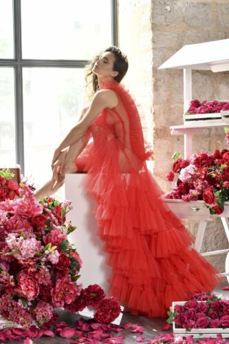 Mannequin portant une robe longue en tulle rouge, style haute couture, posant élégamment parmi des bouquets de fleurs roses et rouges dans un décor lumineux.