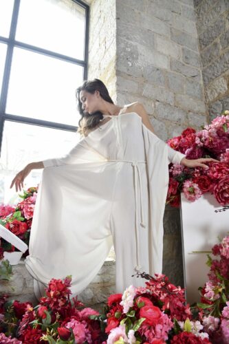 Mannequin portant une robe blanche fluide, style haute couture, posé devant une fenêtre et des fleurs roses, ambiance élégante et sophistiquée.