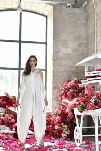 Mannequin portant une élégante robe blanche fluide, posé dans un décor fleuri sophistiqué, près d’une grande fenêtre et d’arrangements floraux roses. Style moderne et raffiné.