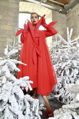 Mannequin portant une robe rouge volumineuse à capuche, au style couture, entouré de sapins enneigés dans un décor hivernal sophistiqué.
