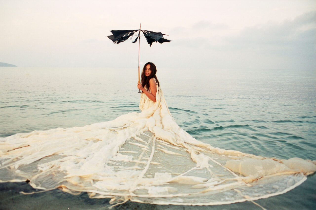 Mannequin debout dans l’eau, portant une longue robe blanche transparente et élégante, tenant un parapluie noir abîmé, atmosphère éthérée et résolument haute couture.