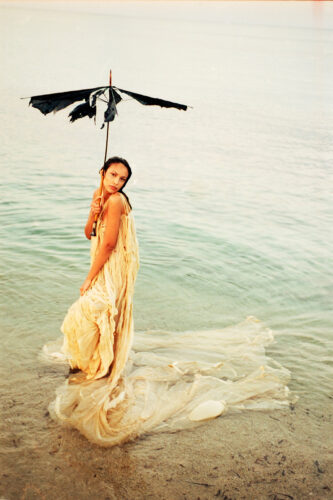 Mannequin portant une robe fluide couleur crème, debout dans l’eau, tenant un parapluie noir abîmé. Scène mêlant mode avant-gardiste et ambiance éthérée.