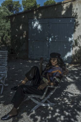 mannequin assis sur une chaise, porte un béret, un pull rayé coloré et un pantalon large, style sophistiqué, exposé à une lumière tamisée extérieure.