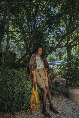 Mannequin habillé d'un short beige taille haute, d'un haut blanc imprimé, d'une veste en fausse fourrure et tenant un sac jaune, posé dans un jardin luxuriant.