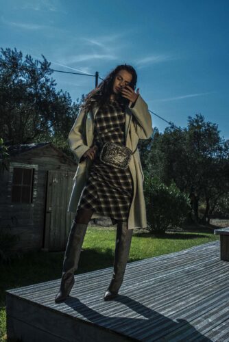 Mannequin posant en extérieur, portant une robe à carreaux, un manteau beige, des bottes hautes et une ceinture, avec une allure élégante et sophistiquée.