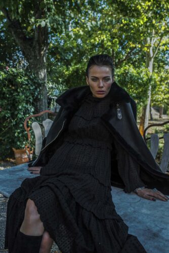 Mannequin vêtu d'une robe noire élégante et d'un manteau sombre, assis sur un banc extérieur, dans un décor naturel sophistiqué.